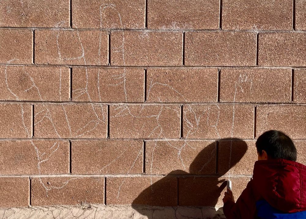 An outline mural of a hand shaping the ASL American manual letter “F”, in yellow chalk on a beige concrete masonry wall, with the boy drawing his own hand on the right side.